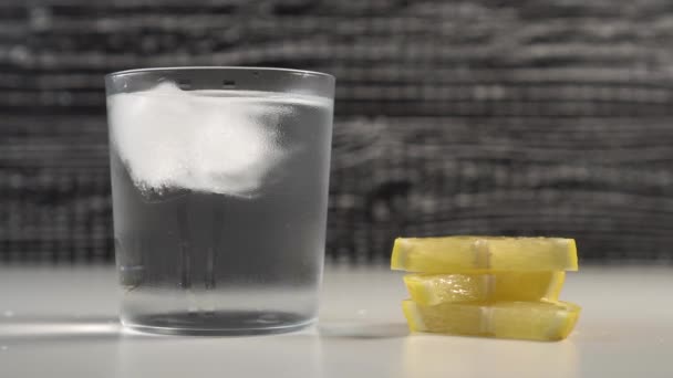 Ice cubes move with water in a misted glass on a black and white background. On a table are slices of fresh lemon - Filmati, video