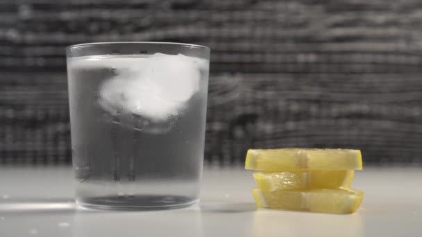 Τα παγάκια κινούνται με νερό σε γυάλινο γυαλί σε μαύρο και άσπρο φόντο. Σε ένα τραπέζι είναι φέτες φρέσκου λεμονιού - Πλάνα, βίντεο