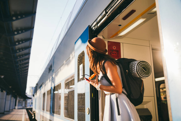 Žena vykukuje vlak. Ženský nádraží. Mladá šťastná žena vytahuje ze dveří vlak a hledá nějakou železniční stanici. Cestování. Portrétní dívka stojící na dveřích vlaku, když dorazila - Fotografie, Obrázek