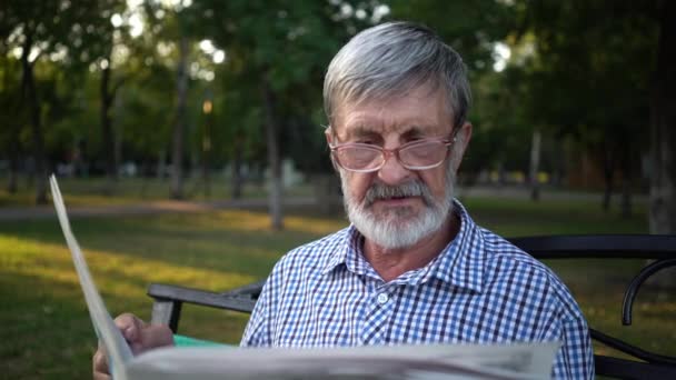 Senior in plaid shirt zit op een bankje in het Park en leest een krant - Video