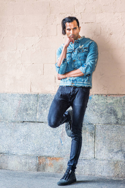 Retrato de Young Modern East Indian American Man em Nova Iorque. Jovem vestindo jaqueta de ganga azul na moda, jeans preto, tênis, de pé contra a parede dobrando a perna, mão tocando o queixo, pensando
 - Foto, Imagem