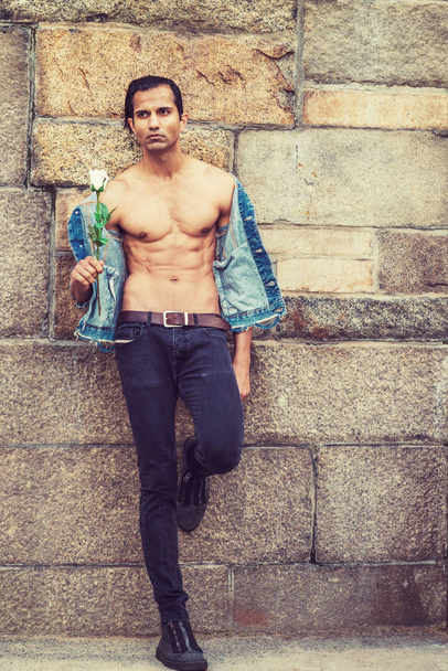 Молодой восточно-индийский американец в синей модной джинсовой куртке, открылся, показывая полуобнаженное сильное тело, черные джинсы, кроссовки, держа белую розу, стоя у каменной стены в Нью-Йорке, ожидая
 - Фото, изображение