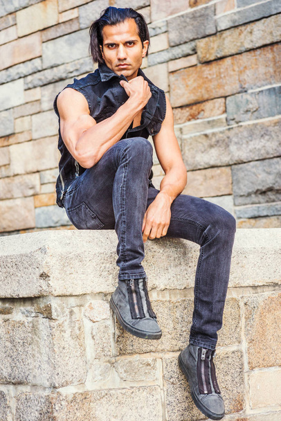 Młody Modern East Indian American Man ubrany w czarny płaszcz bez rękawów, dżinsy, trampki, siedząc na kamiennym ogrodzeniu kamiennym murem w parku w Nowym Jorku, relaks, myślenie - Zdjęcie, obraz