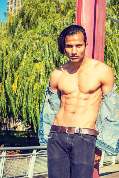 Young East Indian American Man wearing blue Denim jacket, opened, showing half naked strong body, black jeans, standing against red pole at park in New York, serious, intensely looking at you - Foto, Bild