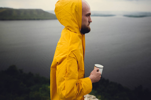 Brutal hombre barbudo en impermeable amarillo sosteniendo taza de metal y trav
 - Foto, imagen