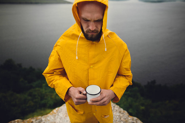 Hipster viajero en impermeable amarillo sosteniendo taza de metal y standi
 - Foto, imagen