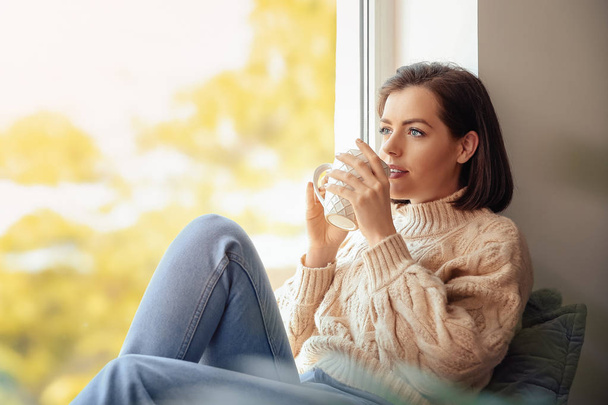 Όμορφη νεαρή γυναίκα πίνοντας καφέ κοντά στο παράθυρο το πρωί - Φωτογραφία, εικόνα