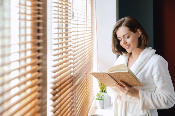 Reggel a szép fiatal nő olvasókönyv közelében ablak - Fotó, kép