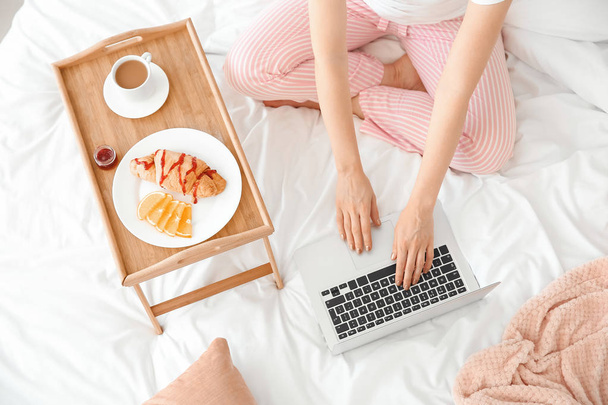 Όμορφη νεαρή γυναίκα με φορητό υπολογιστή που έχει πρωινό στο κρεβάτι - Φωτογραφία, εικόνα