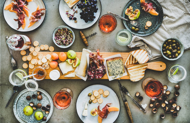 Mid-summer picnic with wine and snacks. Flat-lay of charcuterie and cheese board, rose wine in bottle and glasses, nuts, olives and fruits over concrete table background, top view - Фото, изображение