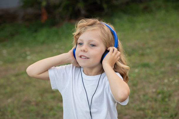 Śmieszne dziecko z długimi włosami słuchanie muzyki z niebieskimi hadphones w parku - Zdjęcie, obraz