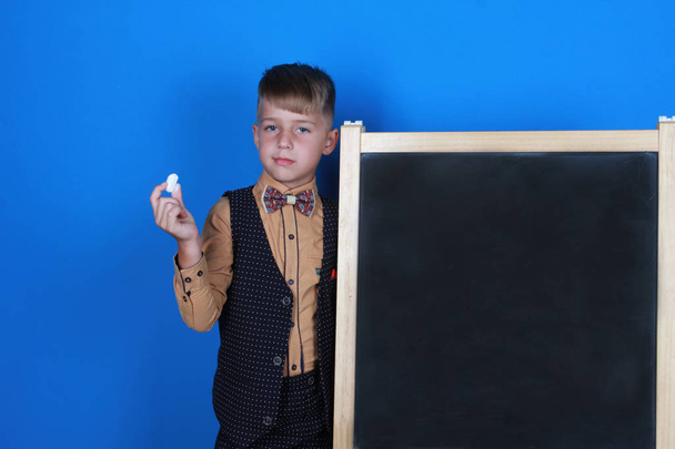 Smart serious schoolboy wearing uniform,  holding chalk and showing blackboard. Boy isolated on blue background. Back to school, education, advertising concept - Fotoğraf, Görsel