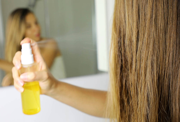 Woman applying oil mask spray on hair in front of a mirror. Haircare concept. Focus on hair. - Fotoğraf, Görsel
