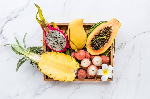Fresh exotic fruits in wooden box on white marble background - sliced papaya, mango, pineapple, dragon fruit, lychee. Mockup, flat lay, overhead. Top view. - Foto, Bild