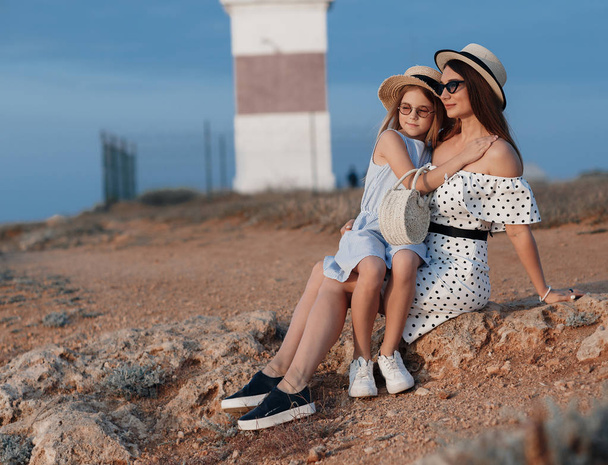 A cheerful child and her mother are enjoying the sunny morning near the water. Portrait of a beautiful brunette mother and her daughter in beautiful dresses on a rocky shore with the sea in the background.Happy family spends vacation at the resort. - Foto, Imagen