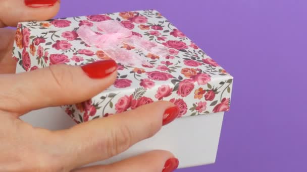 Bir çiçek baskı ve tırnakları mor bir arka plan üzerinde kırmızı oje ile boyanmış kadın elleri tarafından düzenlenen pembe bir yay ile küçük bir hediye kutusu - Video, Çekim