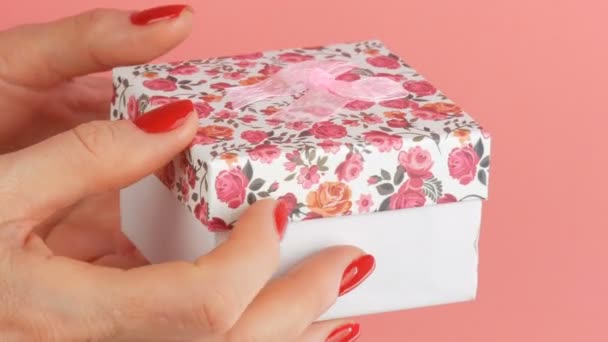 Маленькая подарочная коробка с цветочным шрифтом и розовым бантом, который держат руки женщины, чьи ногти окрашены красным лаком для ногтей на розовом фоне
 - Кадры, видео