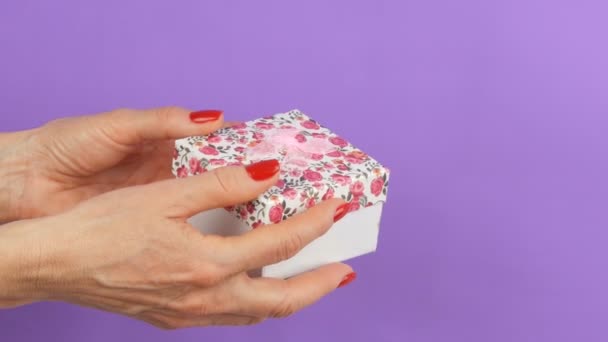 A small gift box with a floral print and a pink bow that is held by the hands of woman whose nails are painted with red nail polish on a purple background - Footage, Video