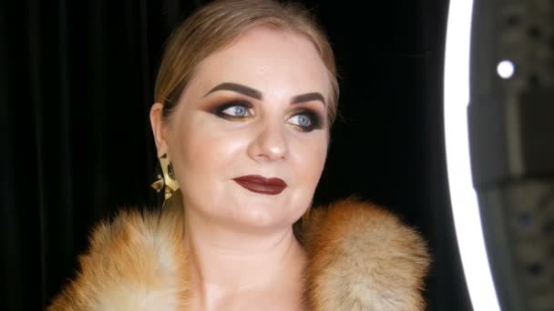 Fat female model with golden makeup and smoky eyes in a coat of fox fur and large golden earrings posing on black background in the studio - Footage, Video