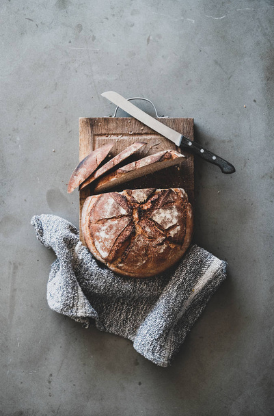 Flat-lay of freshly baked sourdough bread loaf and bread slices on rustic wooden board over grey concrete table background, top view, vertical composition - Foto, afbeelding