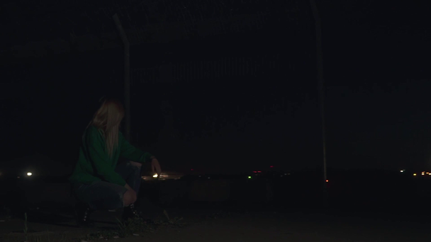 Blond meisje in groene trui hurkt en roken op de voorgrond van het vliegtuig opstijgen. Camera volgt vliegtuig en gaat naar links - Video