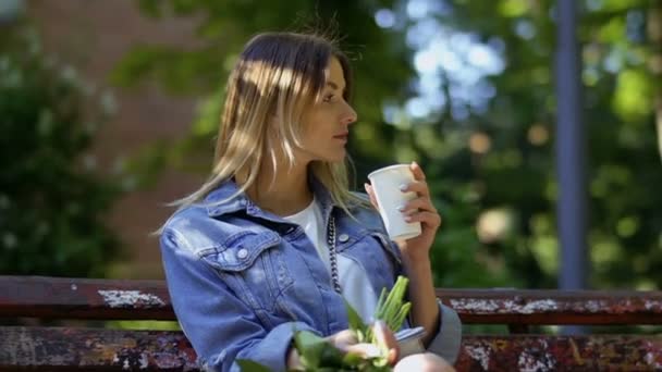 Mladá žena pije kávu z jednorázového šálku, odpočívá na lavičce v letním parku. Hezká blondýna si užívá pití lahodného nápoje. Zpomalený pohyb. - Záběry, video