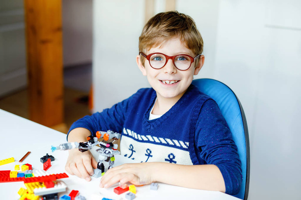 Gözlüklü küçük sarışın çocuk bir sürü renkli plastik blokla oynuyor. Sevimli okul çocuğu robot yaparak ve yaratarak eğleniyor. Yaratıcı eğlence modern teknik ve robotik. - Fotoğraf, Görsel