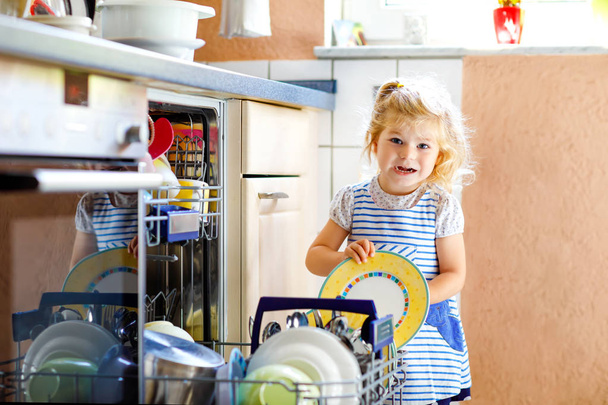 Little adorable cute toddler girl helping to unload dishwasher. Funny happy child standing in the kitchen, holding dishes and putting a bowl on head. Healthy kid at home. Gorgeous helper having fun - Photo, image
