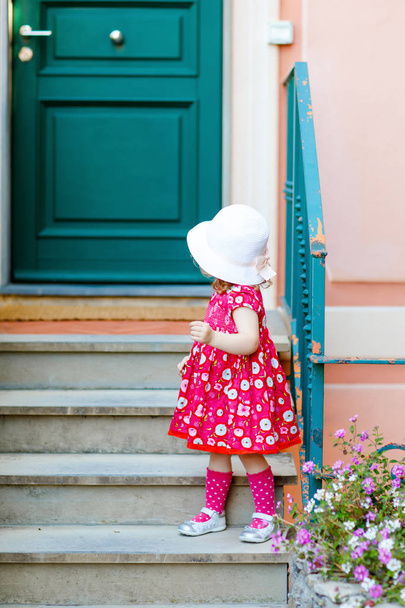 Portrét krásné malé nádherný batole dívka v růžové letní vzhled oblečení, módní šaty, kolena ponožky a klobouk. Šťastné zdravé dítě pózuje před barevným domem. - Fotografie, Obrázek