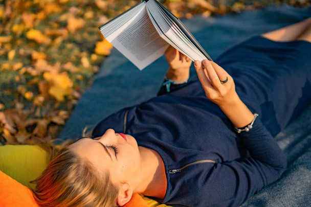 young female in the autumn park. A woman in an autumn park is reading a book, lying on a plaid, autumn reading pleasure. - Photo, Image