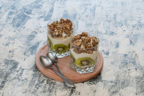 Dessert granola, Greek yogurt, kiwi and banana in two glass cups with spoons on wooden round board, gray concrete. Fitness, figure, body and healthy food - Photo, image