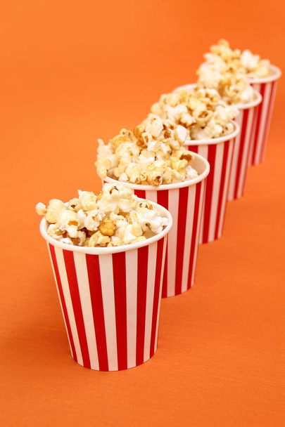 five striped red, white paper cups with popcorn in a row on a bright orange background, close-up, copy space, fast food concept - Foto, imagen