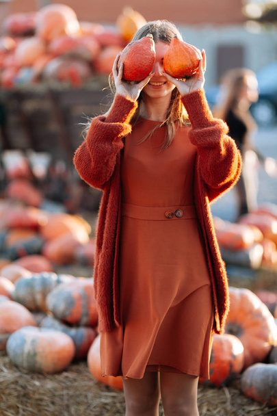 Portrait of happy woman with ripe orange pumpkins in hands close eyes near wagon with orange pumpkin on farmers market in brown sweater, dress. Cozy autumn vibes Halloween, Thanksgiving day - Fotó, kép
