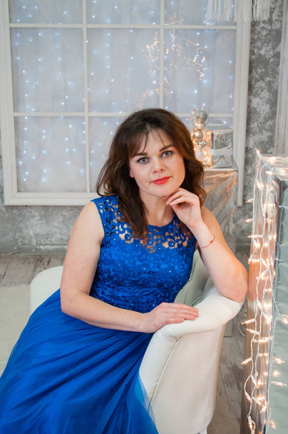 A very beautiful brown-haired woman with curly hair in a blue evening dress is sitting on a business sofa near the New Year's garland. Portrait, vertical format - Photo, Image