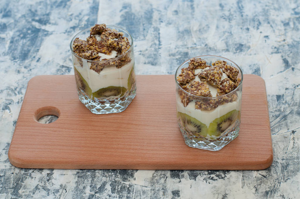 Granola, kiwi, banan i grecki jogurt w dwóch szklanek kubki na drewnianej płycie szary betonu, przestrzeń kopiowania. Fitness, figura, ciało i zdrowa żywność - Zdjęcie, obraz