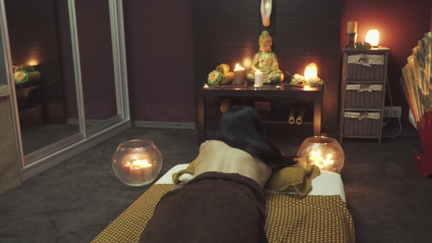 süßes Mädchen liegt in einem Massagebett. Kerzen brennen in der Nähe des Bettes. Auf dem Tisch steht eine Buddha-Statue, Orchideen liegen da und Kerzen brennen. Kamera bewegt sich von links nach rechts. - Filmmaterial, Video