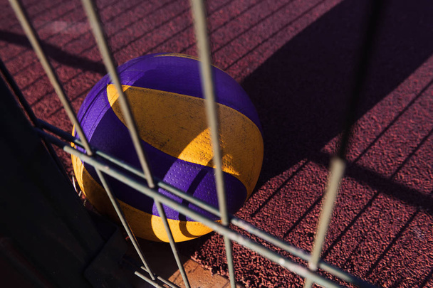 Een eenzame bal leggen op de rode grond na het spel. Verloren volleybal uitrusting. Sportveld bij de zonsondergang met een metalen hek. - Foto, afbeelding