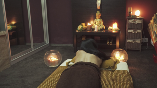 Sweet girl lies in a massage bed. Candles are burning near the bed. On the table there is a Buddha statue, orchids lie and candles are burning. Camera moves from right to left. - 映像、動画