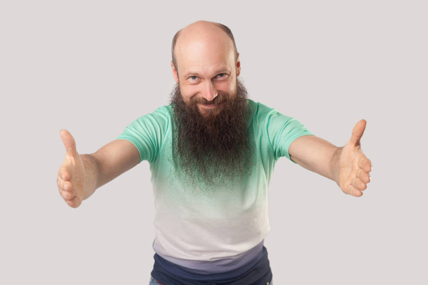 ευτυχισμένος μεσήλικες φαλακρός άνθρωπος με μακρά γενειάδα σε ανοιχτό πράσινο t-shirt στέκεται με ανυψωμένη πλατιά χέρια και κοιτάζοντας με χαμόγελο στο φόντο της κάμερας - Φωτογραφία, εικόνα