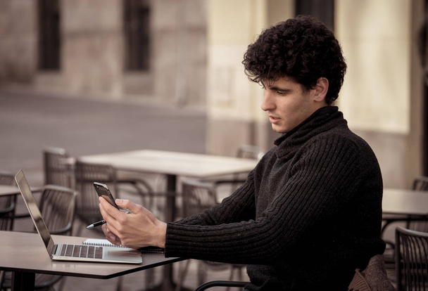 Young man chatting and using smart mobile phone app to talk messaging and taking selfie in a urban european city coffee terrace. In online dating mobile app Internet connections and technology use. - 写真・画像