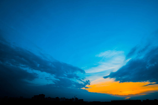 Atmosférico cielo nublado azul detrás de siluetas de edificios de la ciudad. Fondo cobalto y naranja de salida del sol con densas nubes y luz soleada de color amarillo brillante para espacio de copia. Cian cielo por encima de nubes
. - Foto, imagen
