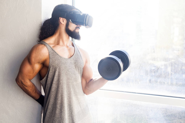 concentrated young adult sportsman with long curly hair holding dumbbell with raised arm and doing exercises for biceps while looking at vr glasses in gym - Fotó, kép