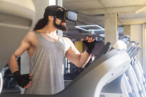 side view of serious young adult sportsman with long curly hair doing exercises on treadmill and looking at vr glasses in gym  - 写真・画像