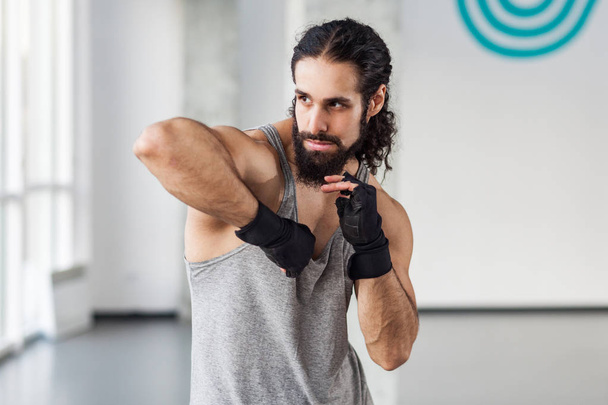 angry muscular young adult man with curly hair in black gloves showing professional kickboxing technique in gym - Фото, изображение