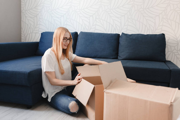 Moving house: Happy woman unpacking box in new home, blonde girl sittng near sofa with boxes - Valokuva, kuva
