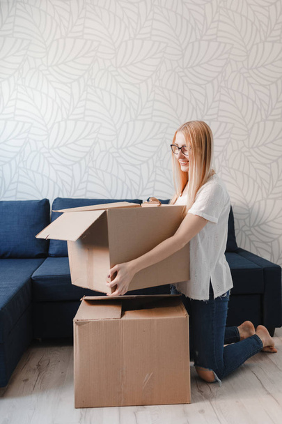 Moving house: Happy woman unpacking box in new home, blonde girl sittng near sofa with boxes - Photo, image