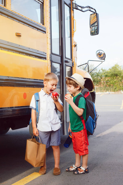 Two funny happy Caucasian boys students kids eating apples near yellow bus on 1 September day. Education and back to school concept. Children pupils ready to learn and study.  - Photo, Image