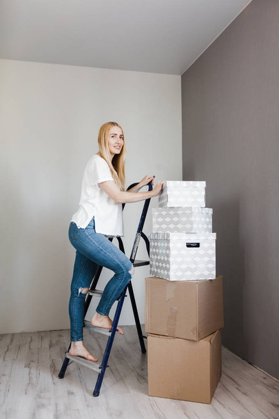 young woman standing on ladder in empty flat with cardboard boxes during relocation at new home - Foto, Imagem