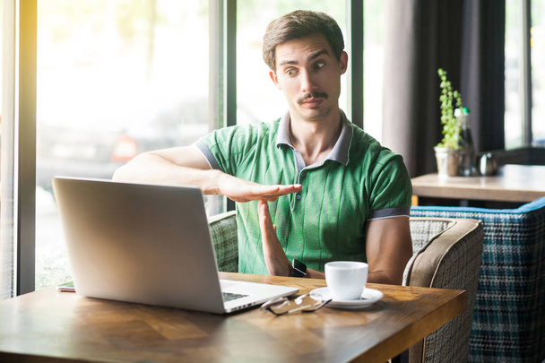 Young businessman in green t-shirt sitting in cafe and looking at laptop screen while having video call and showing time out gesture. business and freelancing concept - 写真・画像