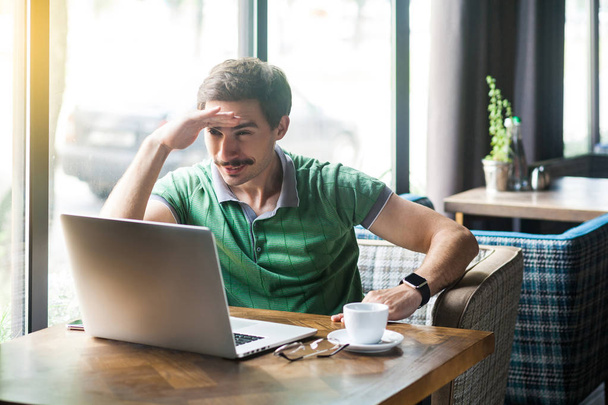 Young attentive businessman in green t-shirt sitting in cafe and working on laptop while holding hand on his forehead. business and freelancing concept  - Foto, immagini